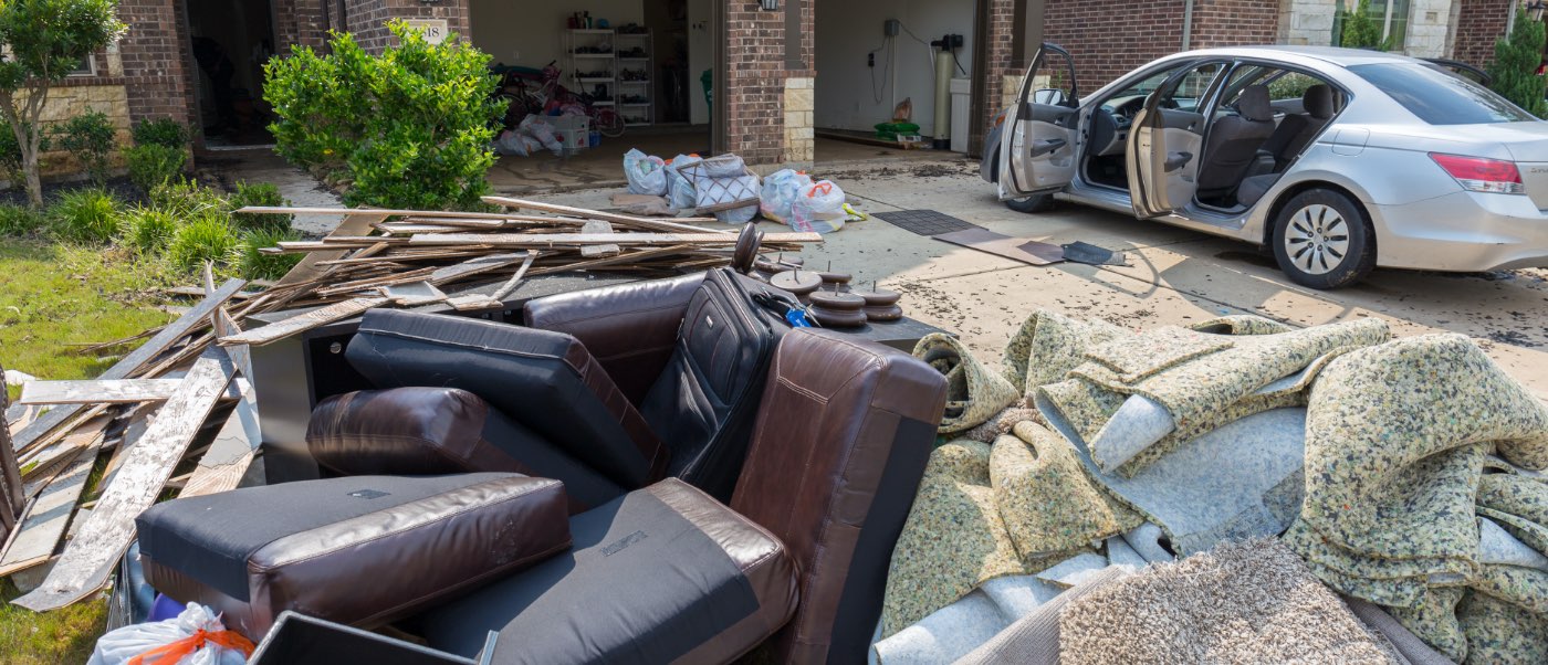 Front yard with discarded carpet and flooring, and other construction remnants with car in driveway with all doors open.